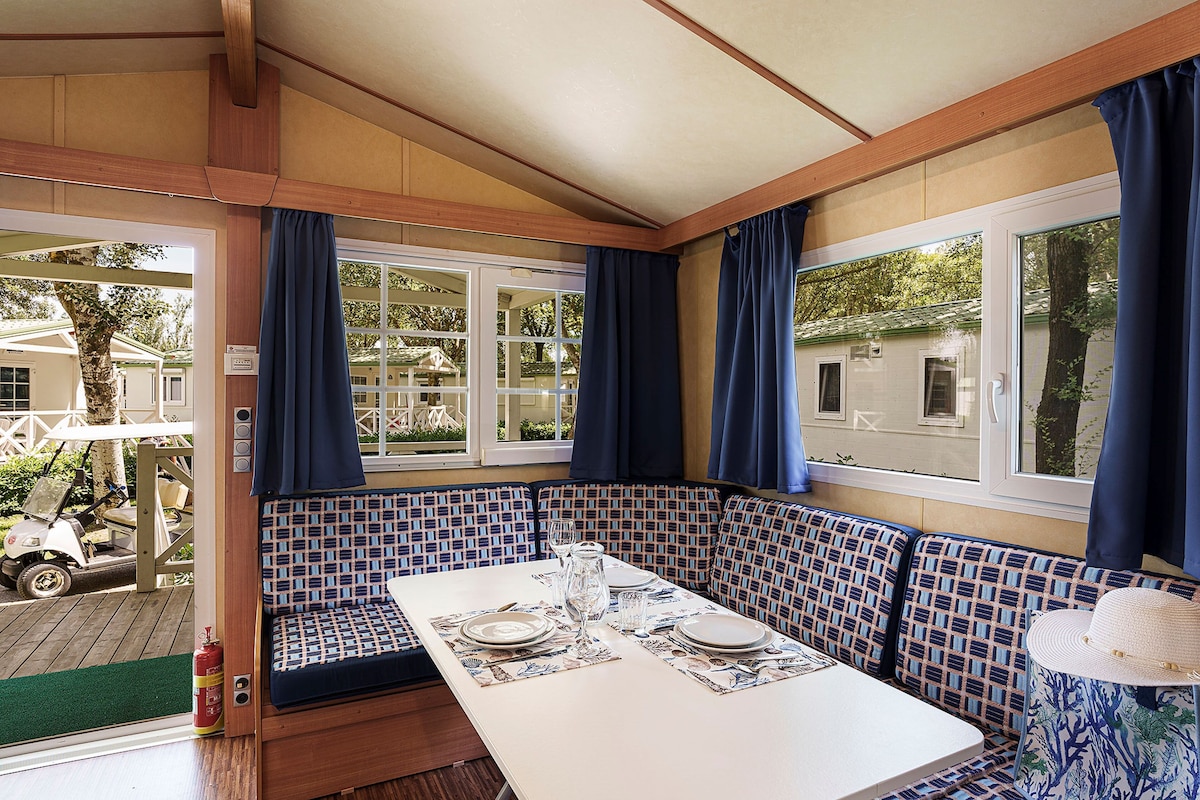 Un coin repas avec une table dressée pour deux personnes, un banc agrémenté d’un coussin à motifs et une vue sur l’extérieur grâce à de grandes fenêtres.