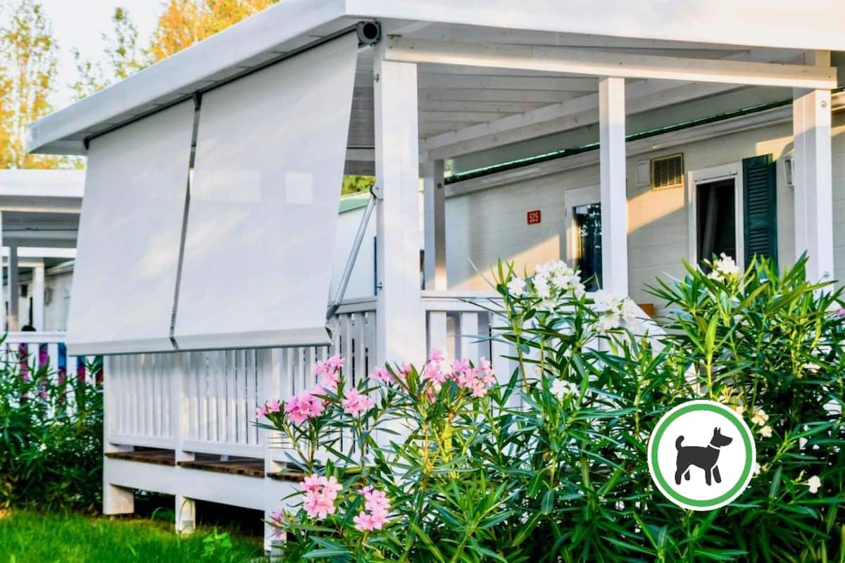 A white covered outdoor area with a wooden deck, surrounded by green foliage and pink flowers.