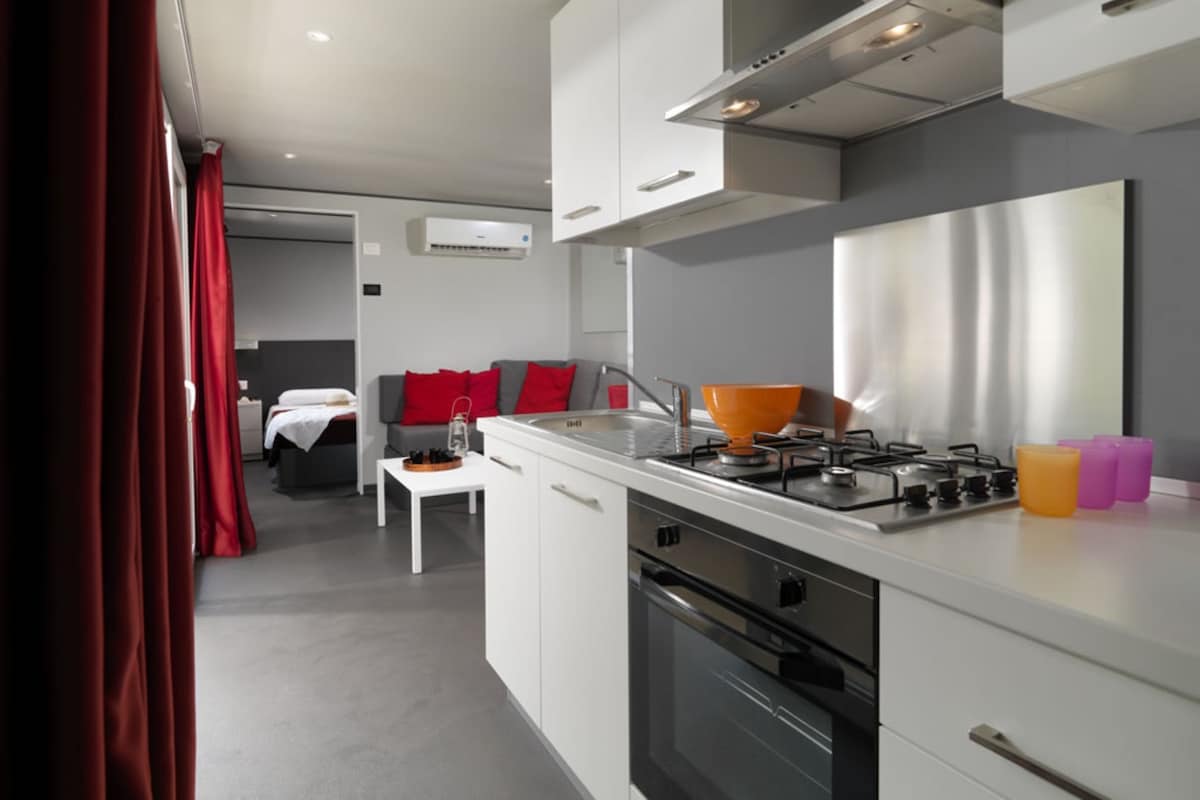 Une cuisine moderne avec des armoires blanches, une cuisinière à gaz et un four encastré. On y trouve une petite table à manger avec une chaise, et un rideau rouge est visible sur le côté gauche.