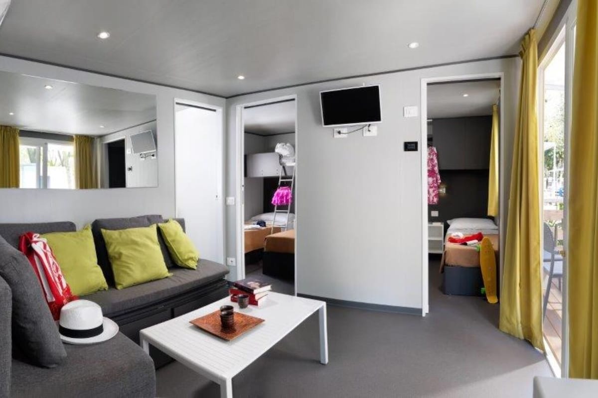 Une chambre d’hôtel moderne avec un canapé gris, une table basse blanche et un lit agrémenté de rideaux jaunes.