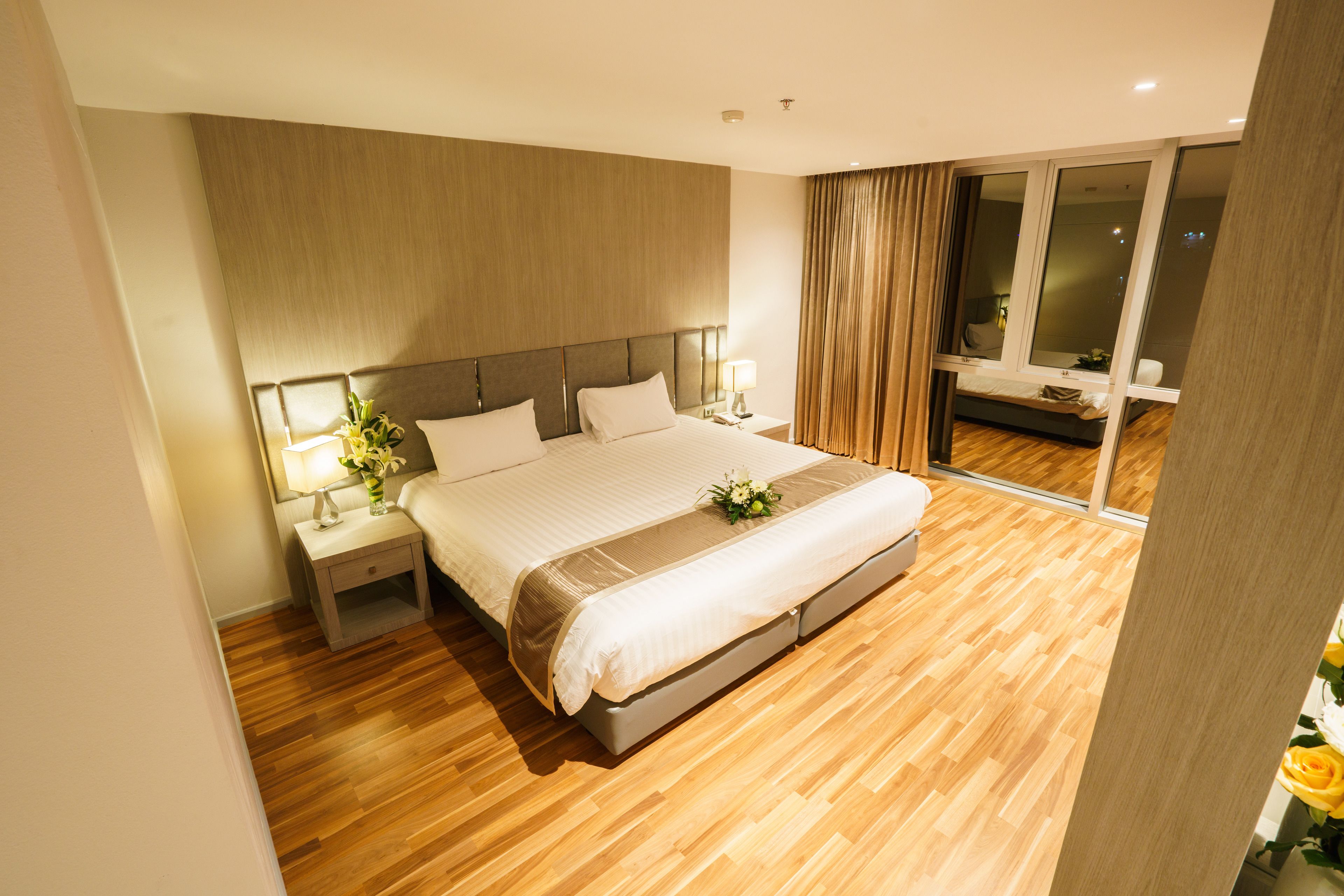 A modern hotel room with a large bed, wooden flooring, a nightstand with a lamp, and a view of the city through a large window.