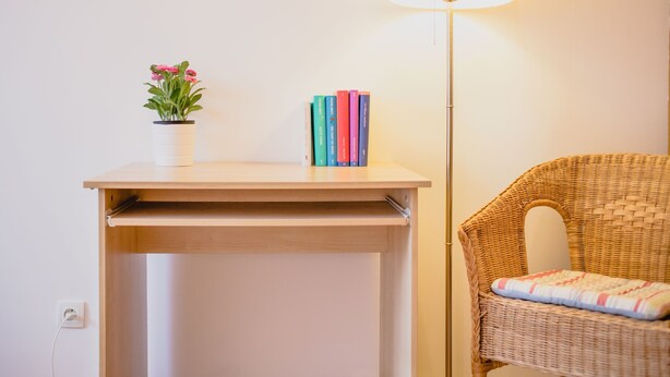 A small wooden table with a potted plant and a stack of colorful books. A wicker chair with a cushion is next to a floor lamp.