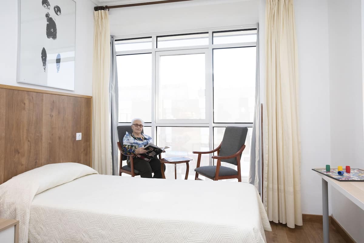 A hotel room with a bed, a window with curtains, a small table with colorful objects, and a chair.
