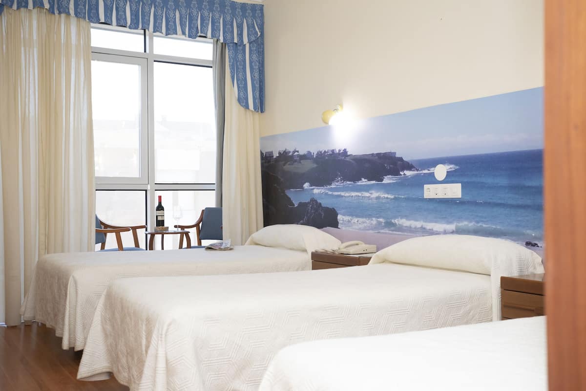 A hotel room with two beds, a window with curtains, a table with a chair, and a wall mural of a coastal scene.
