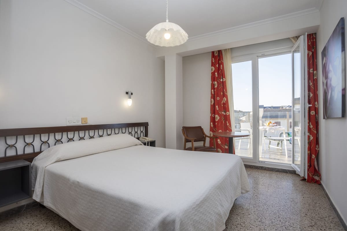 A hotel room with a bed, a chair, a small table, a window with curtains, and a balcony with outdoor furniture.