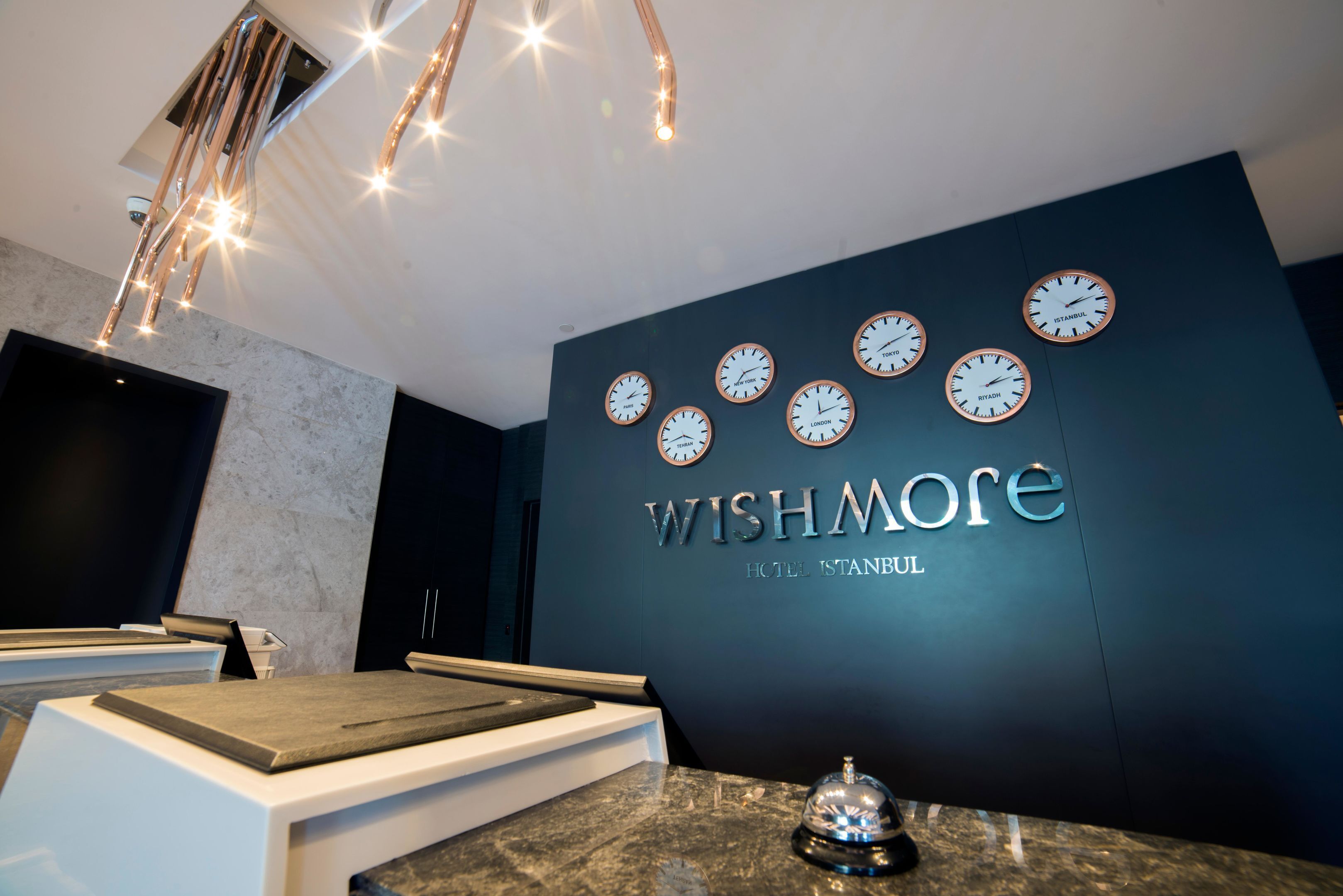 Zona de recepción moderna de un hotel, con una pared azul oscuro, varios relojes mostrando diferentes horas y un mostrador de recepción con encimera de madera.
