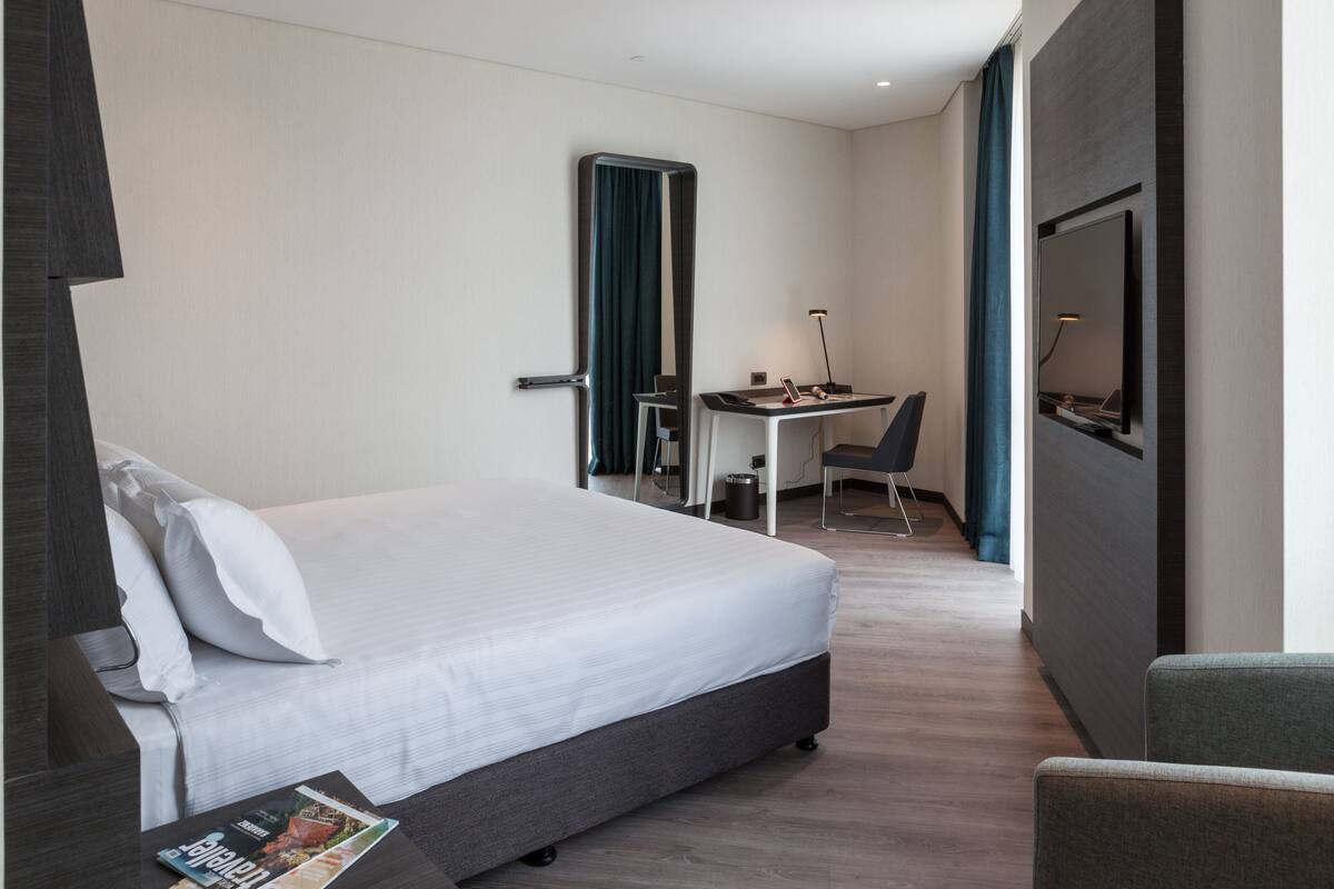 A hotel room with a bed, a desk with a chair, a TV, and a magazine on the nightstand.