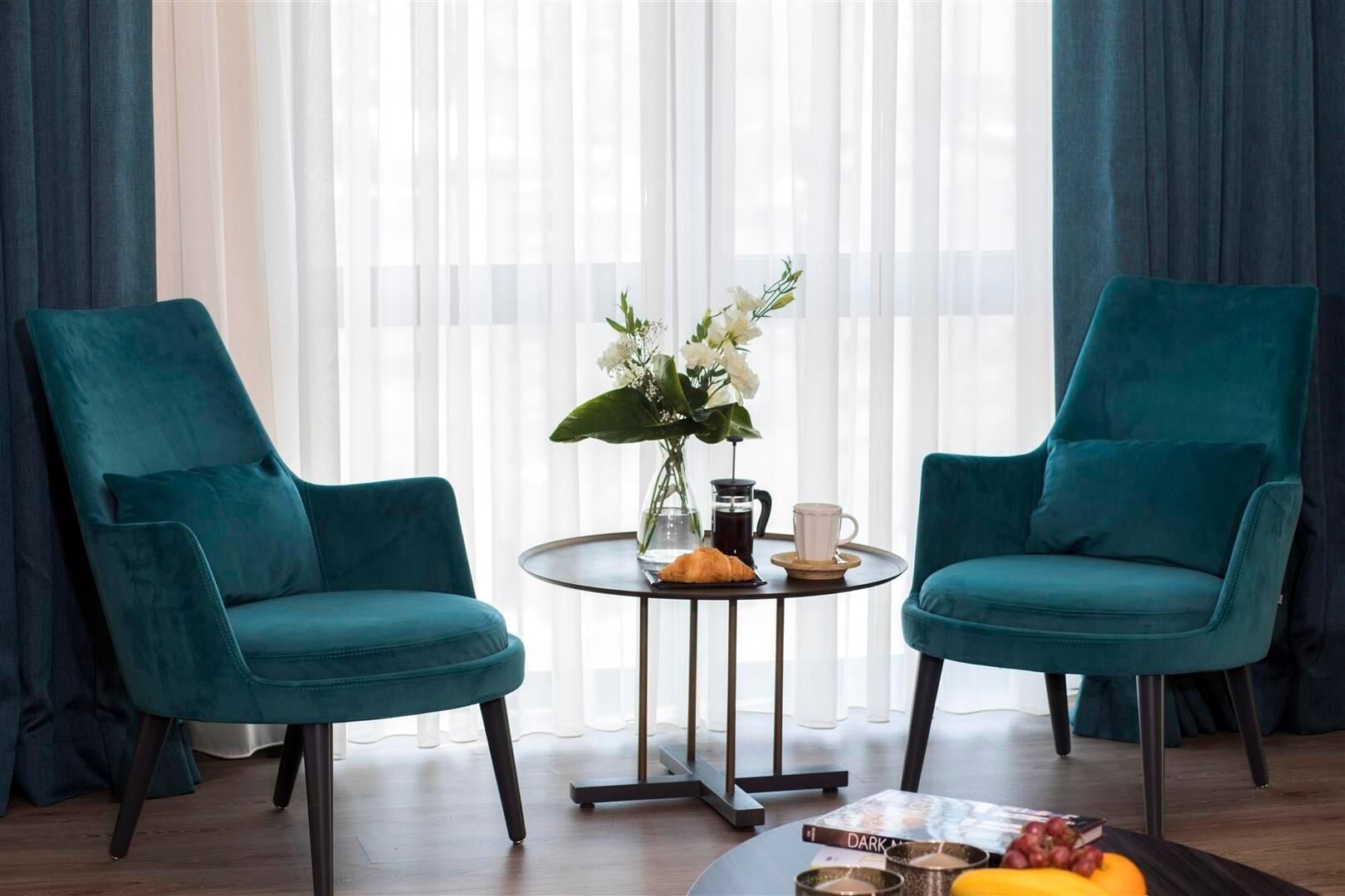 A room with two teal armchairs, a small round table with a coffee pot, cup, and flowers, and a book on the table.