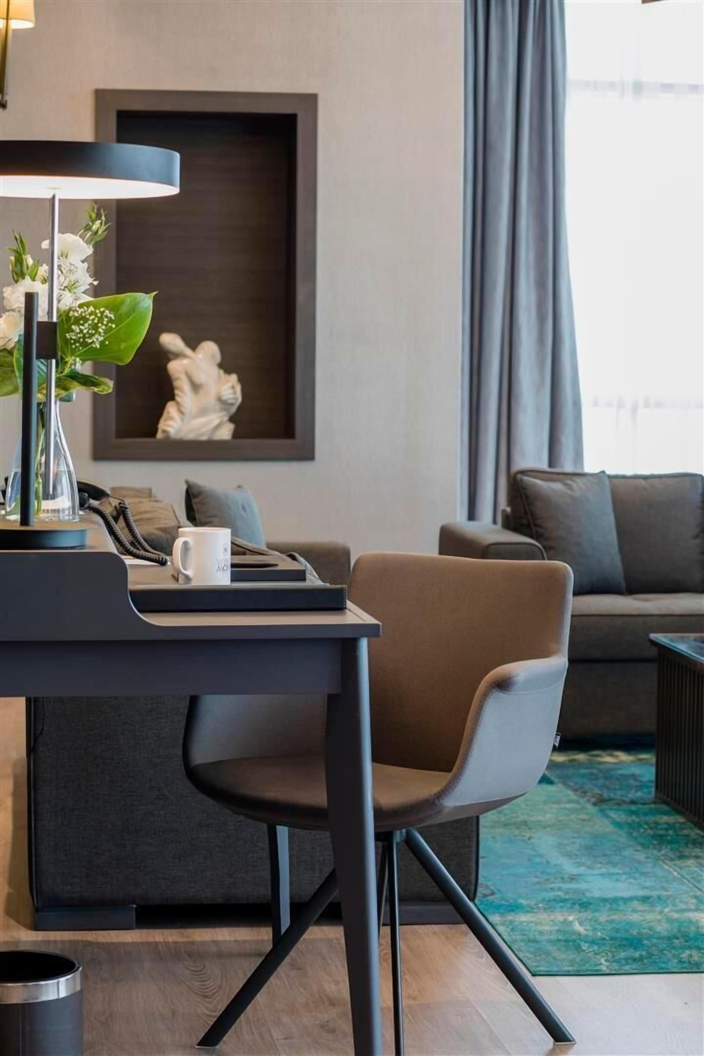 Ein modernes Wohnzimmer mit einem dunklen Sofa, einem Couchtisch und einem Schreibtisch mit Lampe und Blumenvase.