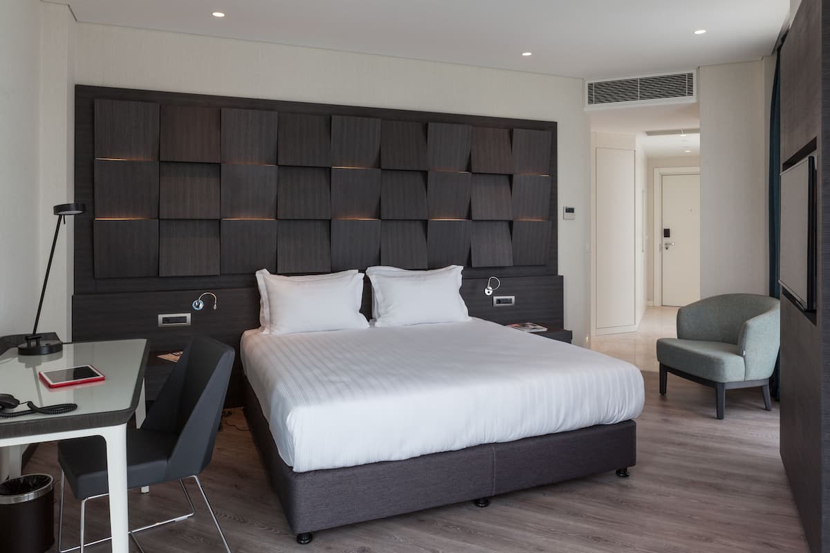 A modern hotel room with a large bed, a desk with a chair, a chair, and a bedside table.