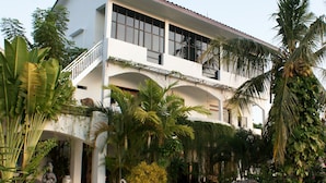 Una casa de dos pisos con balcón, rodeada de plantas tropicales y una piscina.