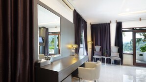 Habitación de hotel moderna con un espejo grande, una tocador con lámpara, una silla blanca y vistas a la vegetación por la ventana.