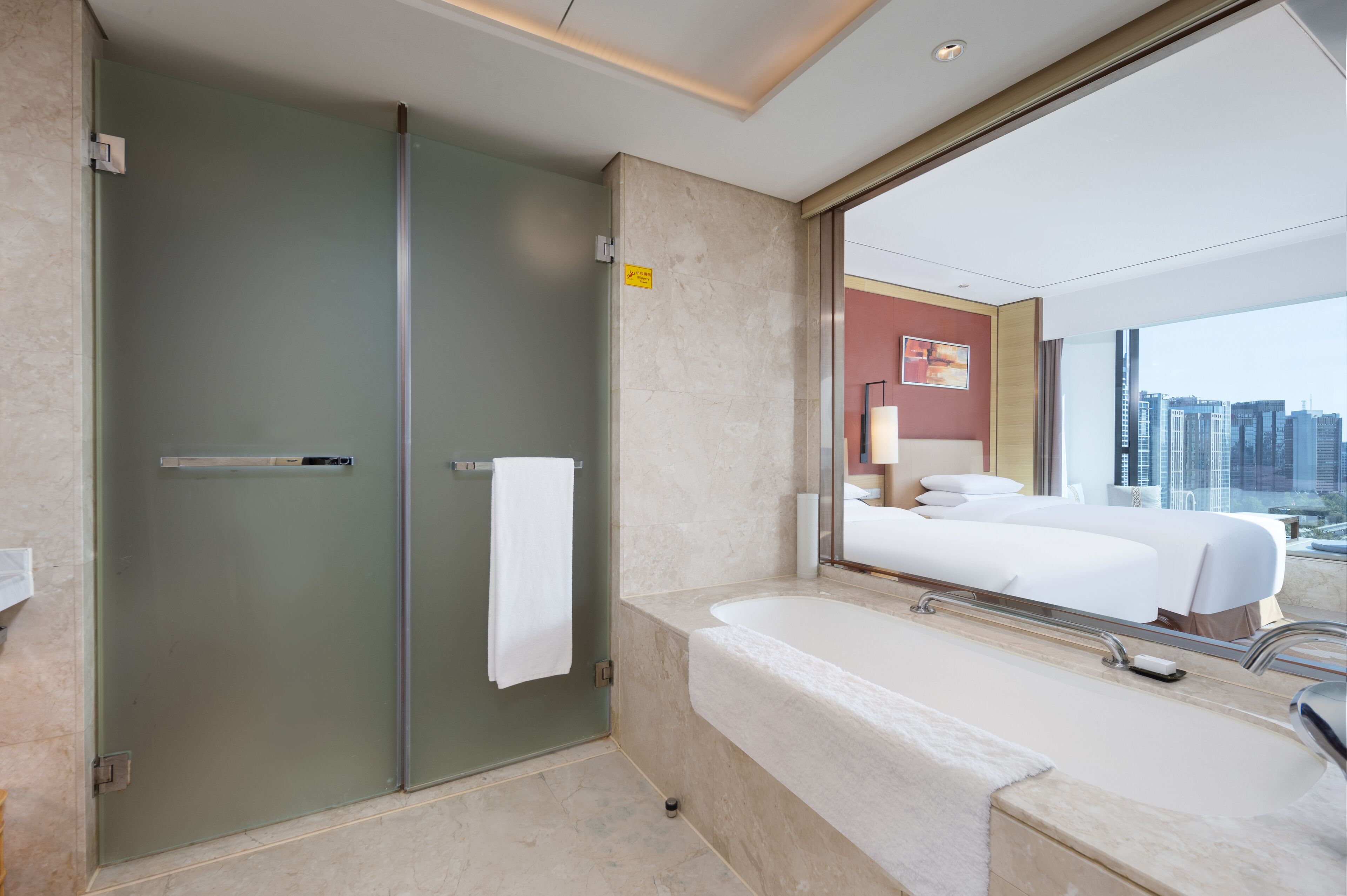 A modern hotel bathroom with a glass shower enclosure, a large bathtub, and a city view through a large window.