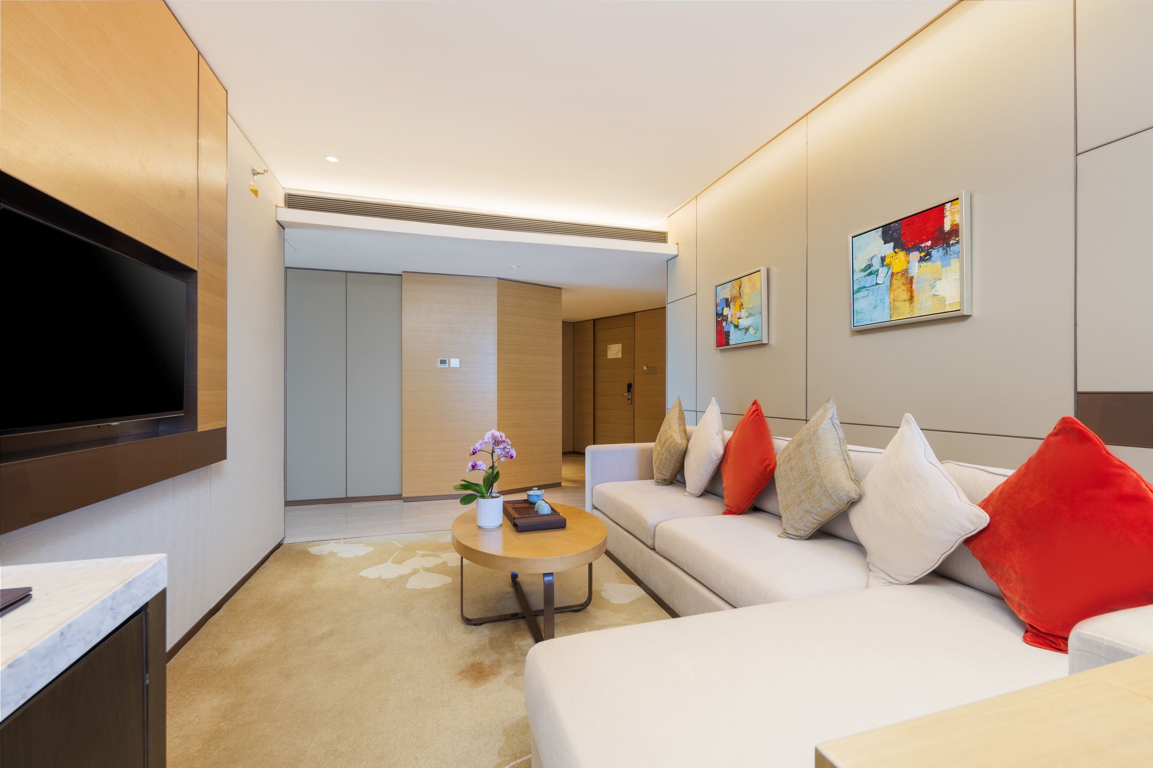 Habitació d'hotel moderna amb televisió de pantalla plana, un sofà amb coixinets vermells i beix, una taula de cafè rodona amb un ram de flors i una obra d'art abstracta a la paret.