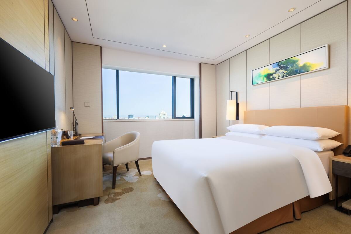 Ein Hotelzimmer mit einem großen Bett, einem Schreibtisch, einem Stuhl, einem Fernseher und einem Fenster mit Blick auf die Stadt.