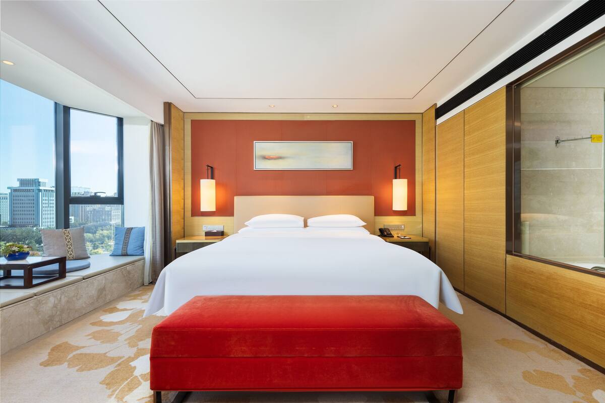 Ein modernes Hotelzimmer mit einem großen Bett, einem roten Pouf, Blick auf die Stadt und einem Bild an der Wand.
