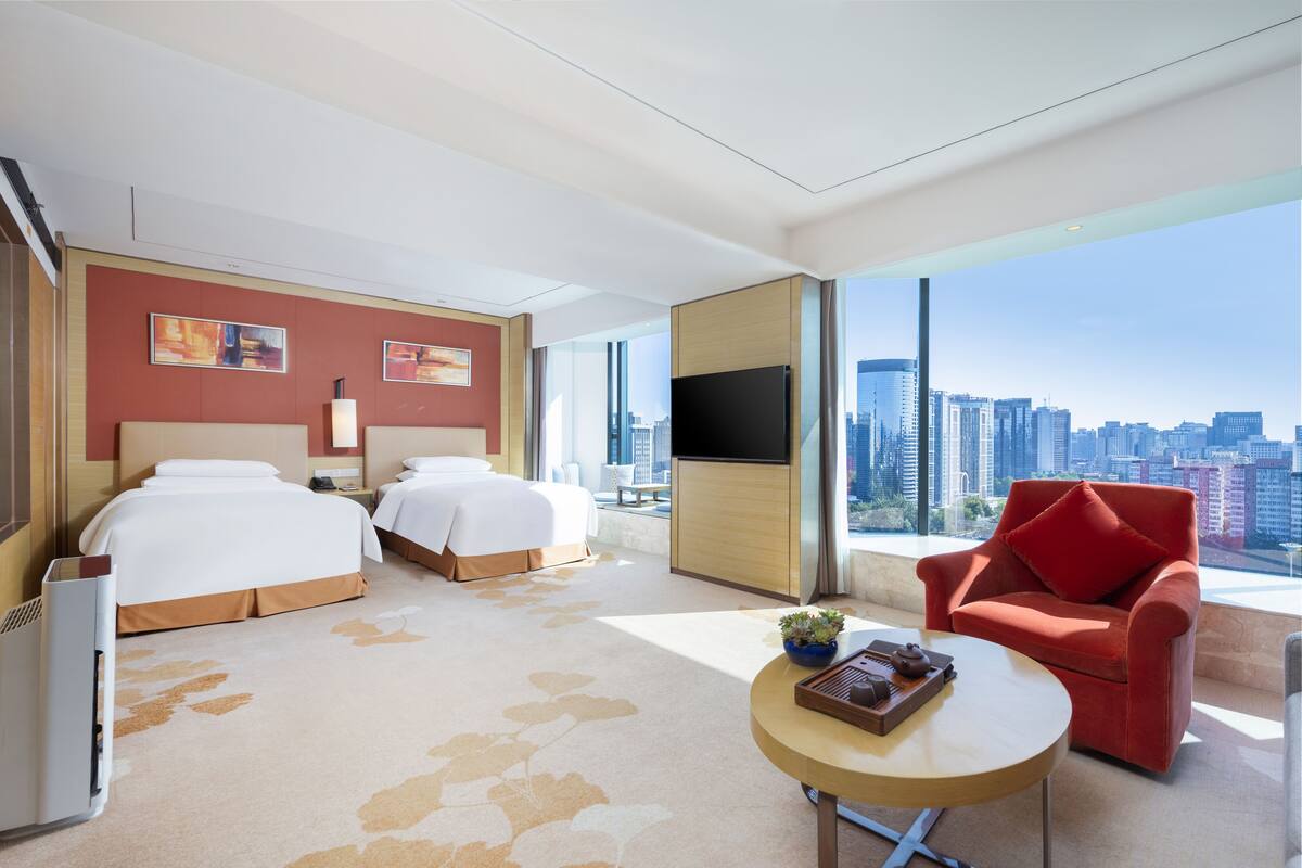 Ein Hotelzimmer mit zwei Betten, einem roten Sessel, einem runden Tisch, einem Fernseher und Blick auf die Stadt.