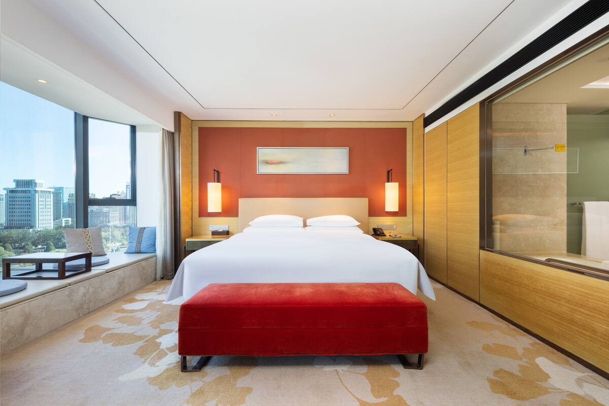 Ein modernes Hotelzimmer mit einem großen Bett, einer roten Bank, Blick auf die Stadt und einer Duschkabine aus Glas.