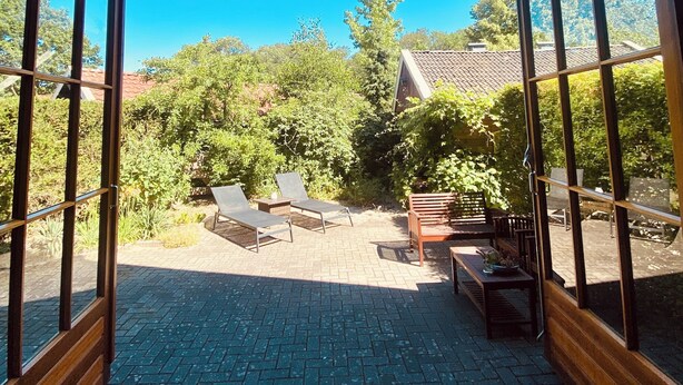 Eine offene Terrasse mit Backsteinböden, Gartenmöbeln und Blick auf einen Garten sowie ein Haus durch große Fenster.