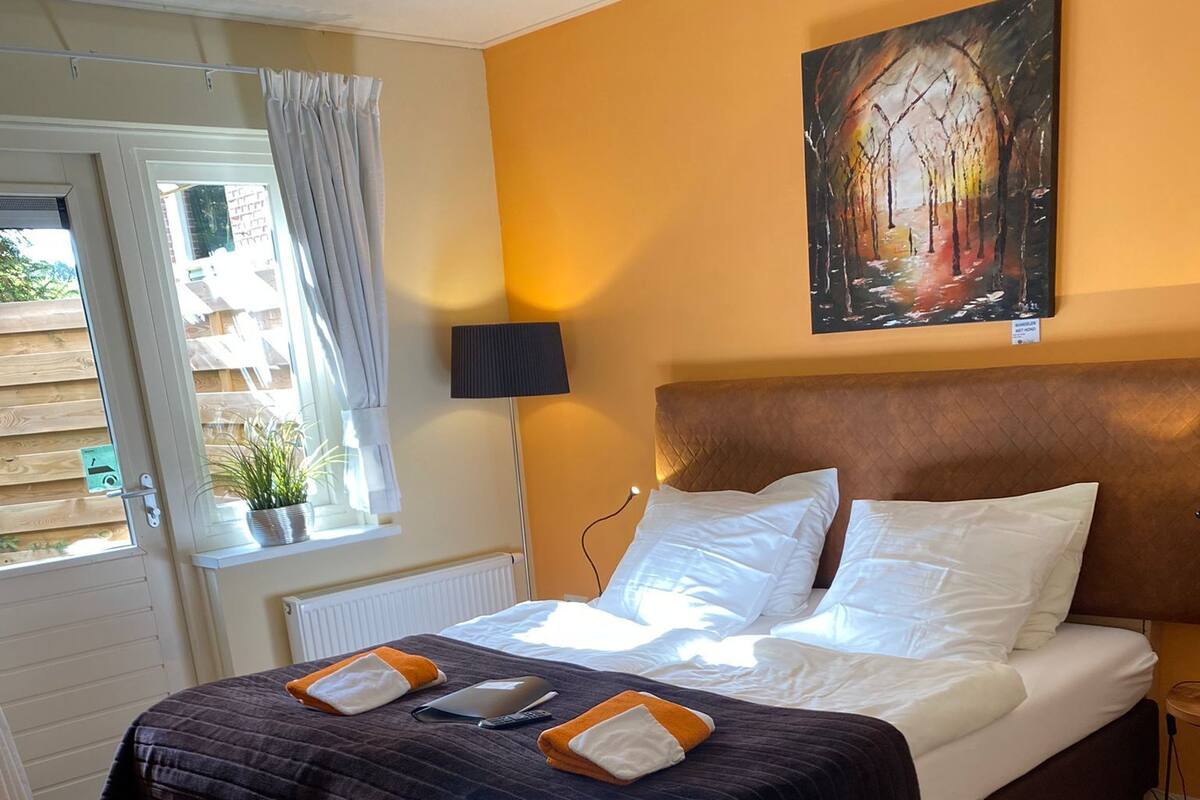 Quarto de hotel com cama, mesa de cabeceira, luminária, um quadro na parede e janela com cortinas.