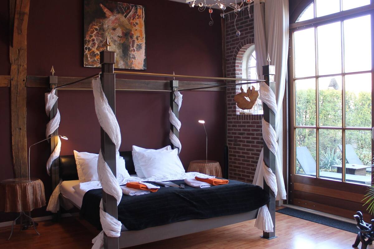Uma cama com dossel com lençóis brancos, um quadro de uma girafa na parede e vista para o verde através de uma janela grande.