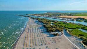 Luftaufnahme einer Küstenlandschaft mit einem langen Sandstrand, Reihen von Strandsonnenschirmen und einer Marina mit Booten.