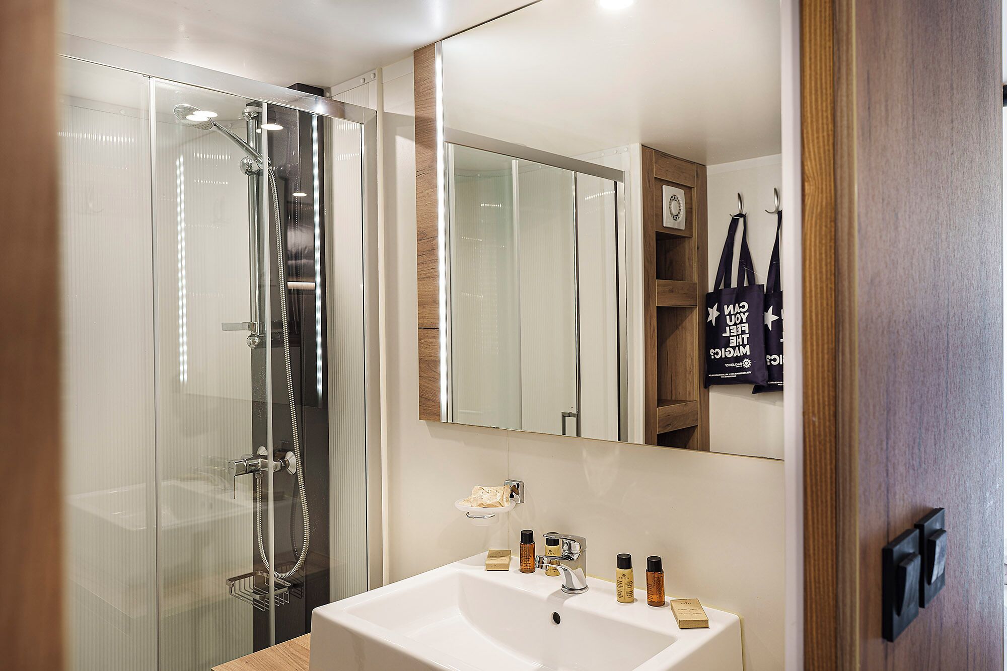 A modern bathroom with a glass-enclosed shower, a white sink with toiletries, and a mirror reflecting the room.