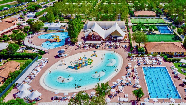 Eine Vogelperspektive zeigt ein Resort mit mehreren Schwimmbecken, einem Strandbereich und einem großen Gebäude mit markanter Dachkonstruktion.