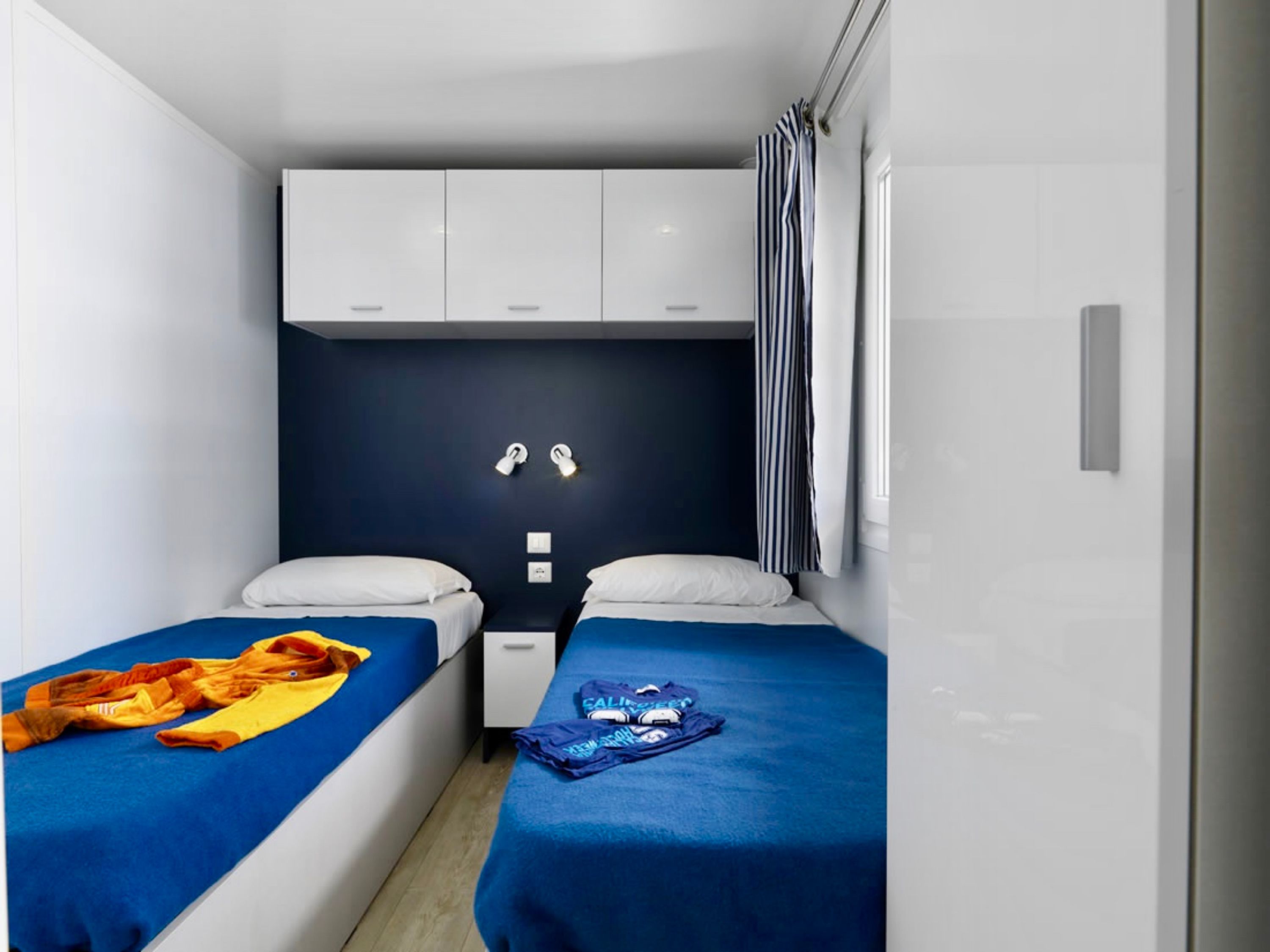Une chambre d’hôtel de taille réduite, avec deux lits simples, un petit bureau et un mur de couleur sombre.