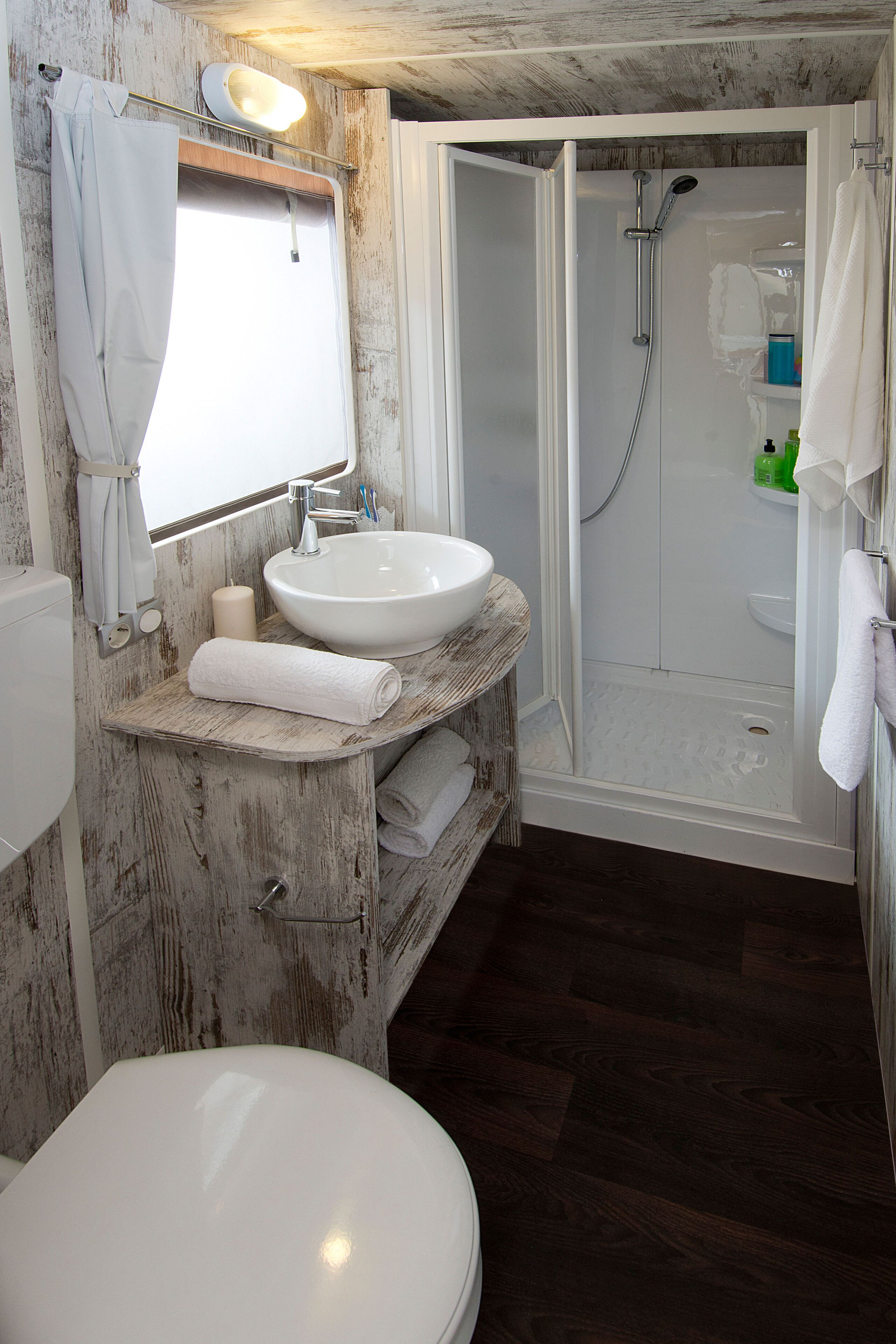 Un baño con lavamanos blanco, espejo e inodoro. Cuenta con una zona de ducha con puerta de vidrio y un perchero.