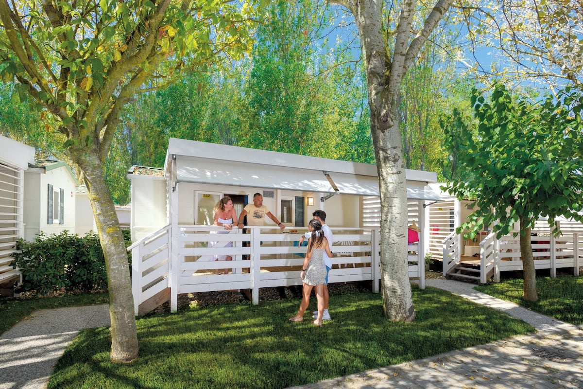 A row of white mobile homes with a covered outdoor area, surrounded by trees and a well-maintained lawn.