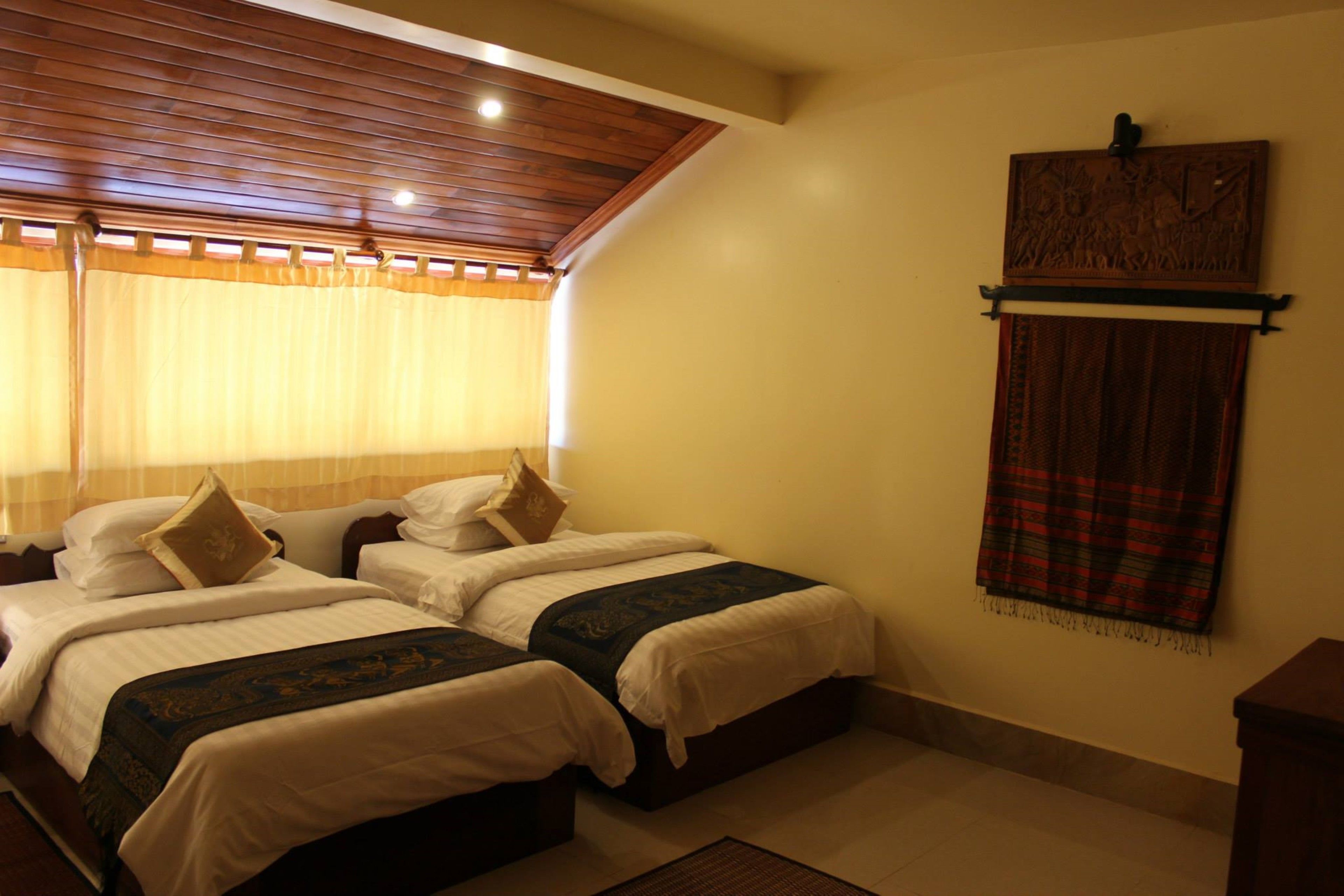 A bedroom with two beds, a wooden ceiling, a wall-mounted shelf, and a window with curtains.
