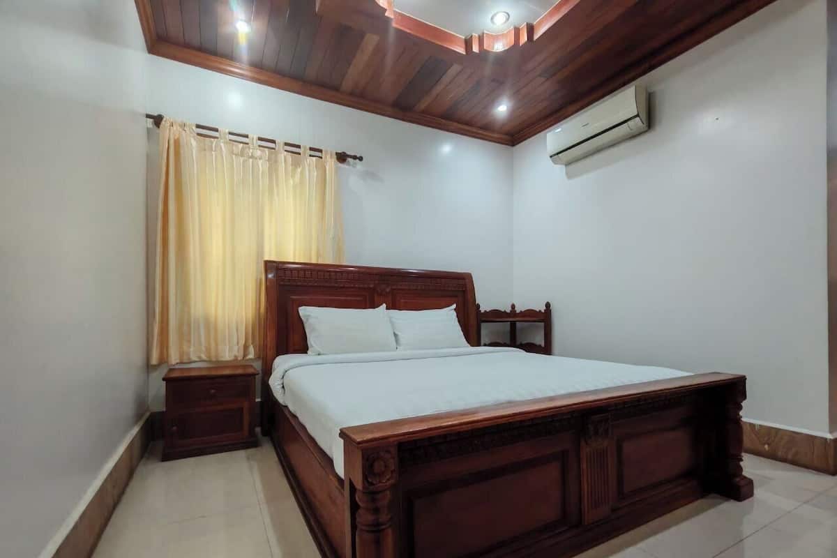 Une chambre avec un lit en bois, une table de chevet, une fenêtre avec des rideaux et un appareil de climatisation fixé au mur.