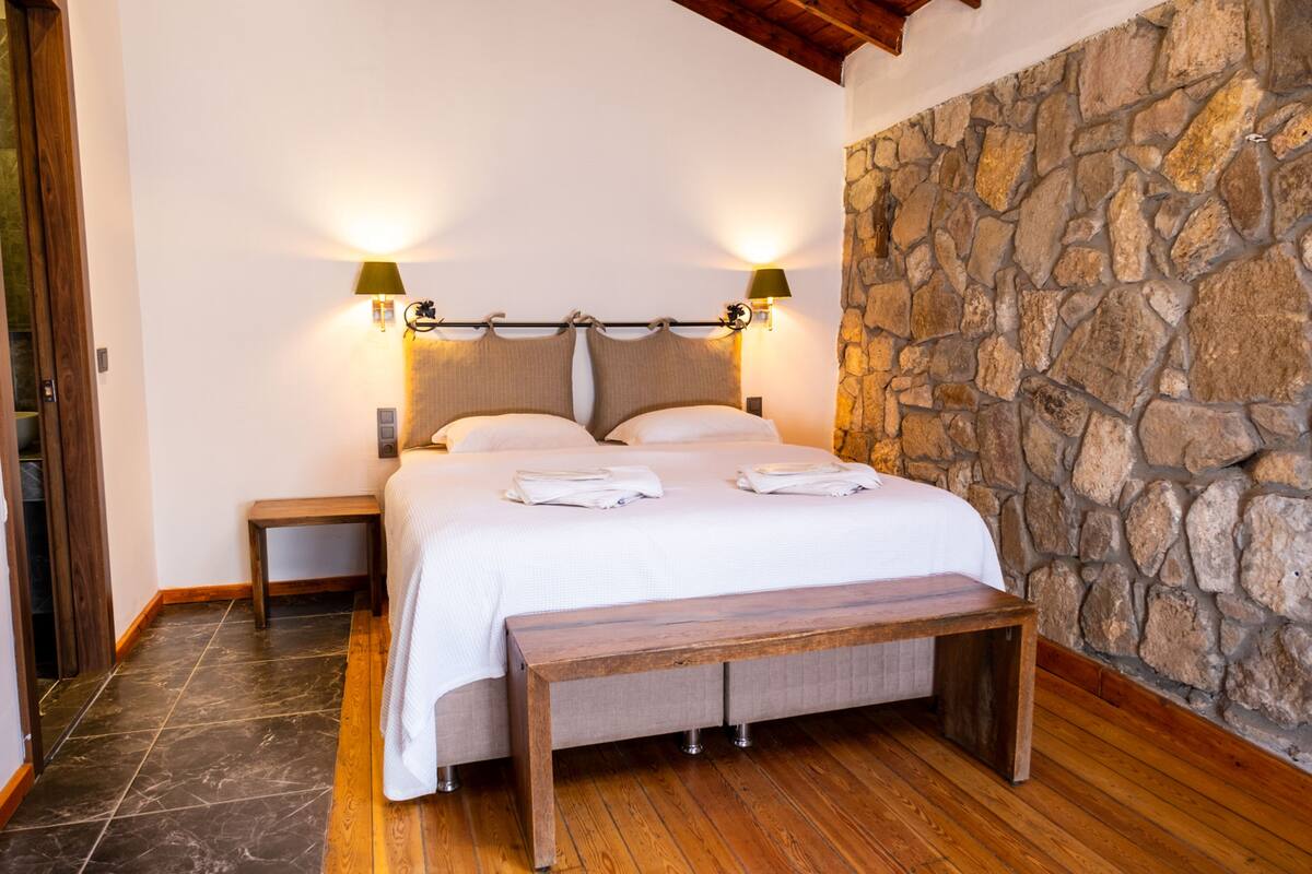 Un dormitorio con pared de piedra, suelo de madera, una cama con ropa de cama blanca, dos mesitas de noche y dos lámparas de pared.