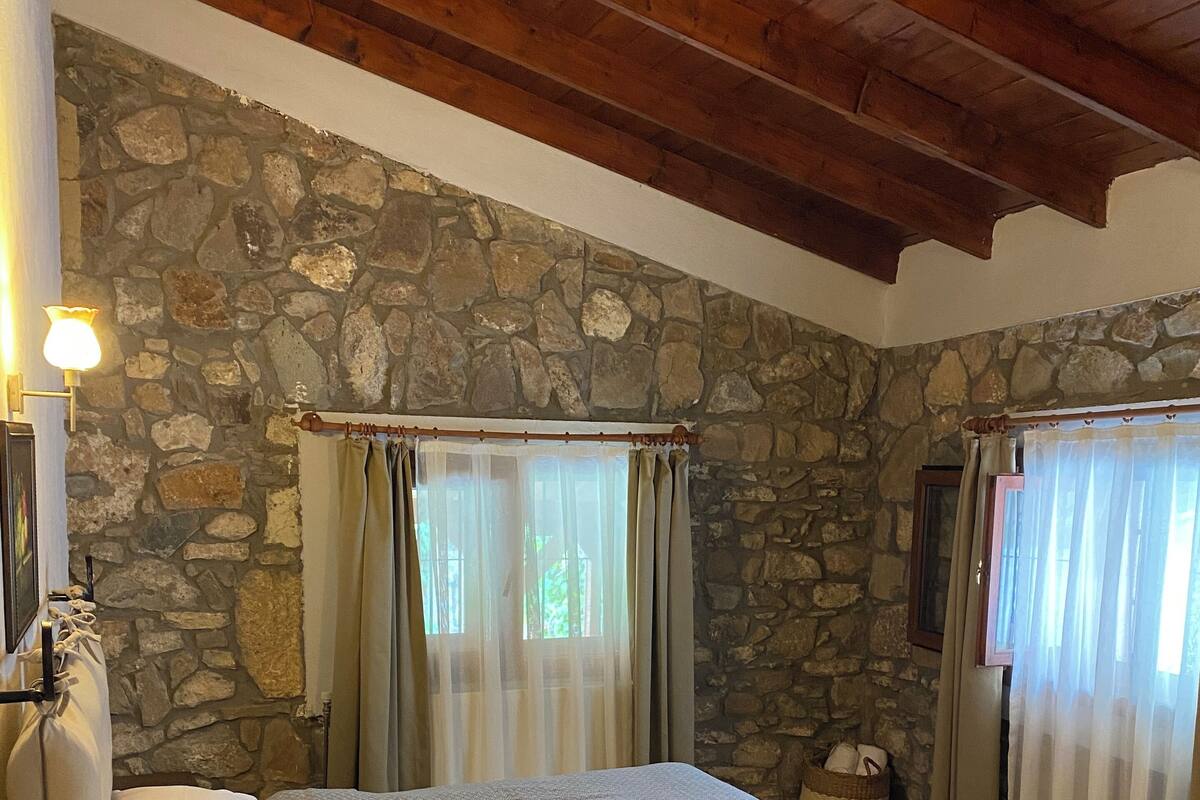 Un dormitorio con pared de piedra, vigas de madera en el techo, una cama con colcha estampada y una mesita de noche.