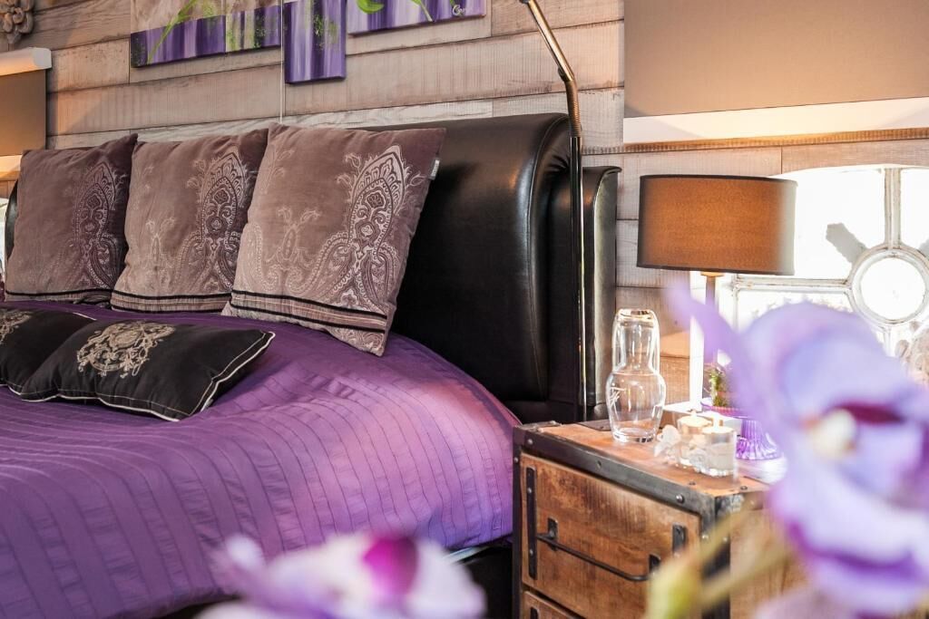 A bed with a purple quilt and decorative pillows, a wooden nightstand with a lamp, and a window with a view of the sky.