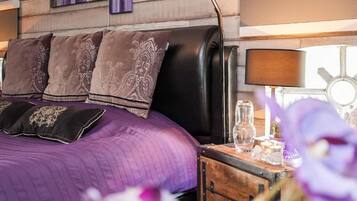 A bed with a purple quilt and decorative pillows, a wooden nightstand with a lamp, and a window with a view of the sky.