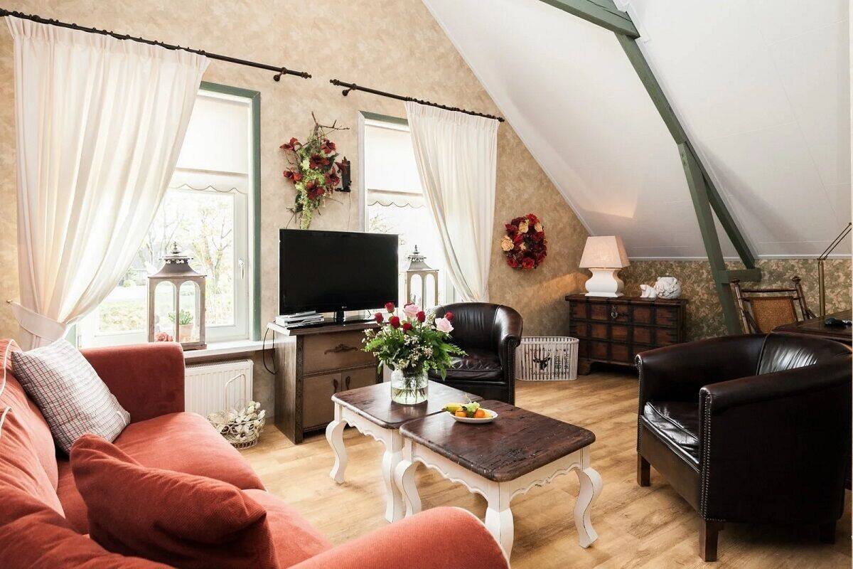 Un salon chaleureux avec un canapé rouge, un fauteuil en cuir noir, une table basse en bois ornée d’un vase de fleurs, un téléviseur et une guirlande décorative accrochée au mur.