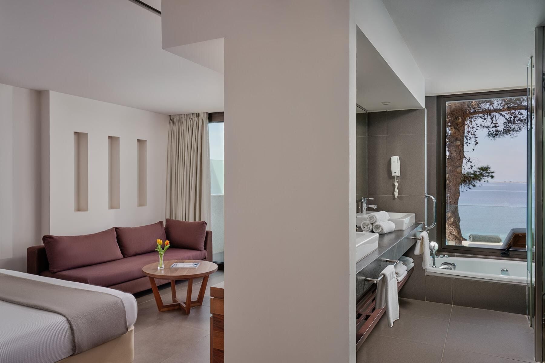A modern hotel room with a sofa, a small round table, and a view of the sea through a large window.