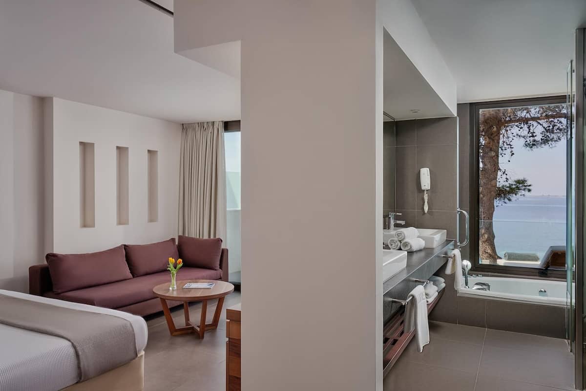 A modern hotel room with a sofa, a small round table, and a view of the sea through a large window.