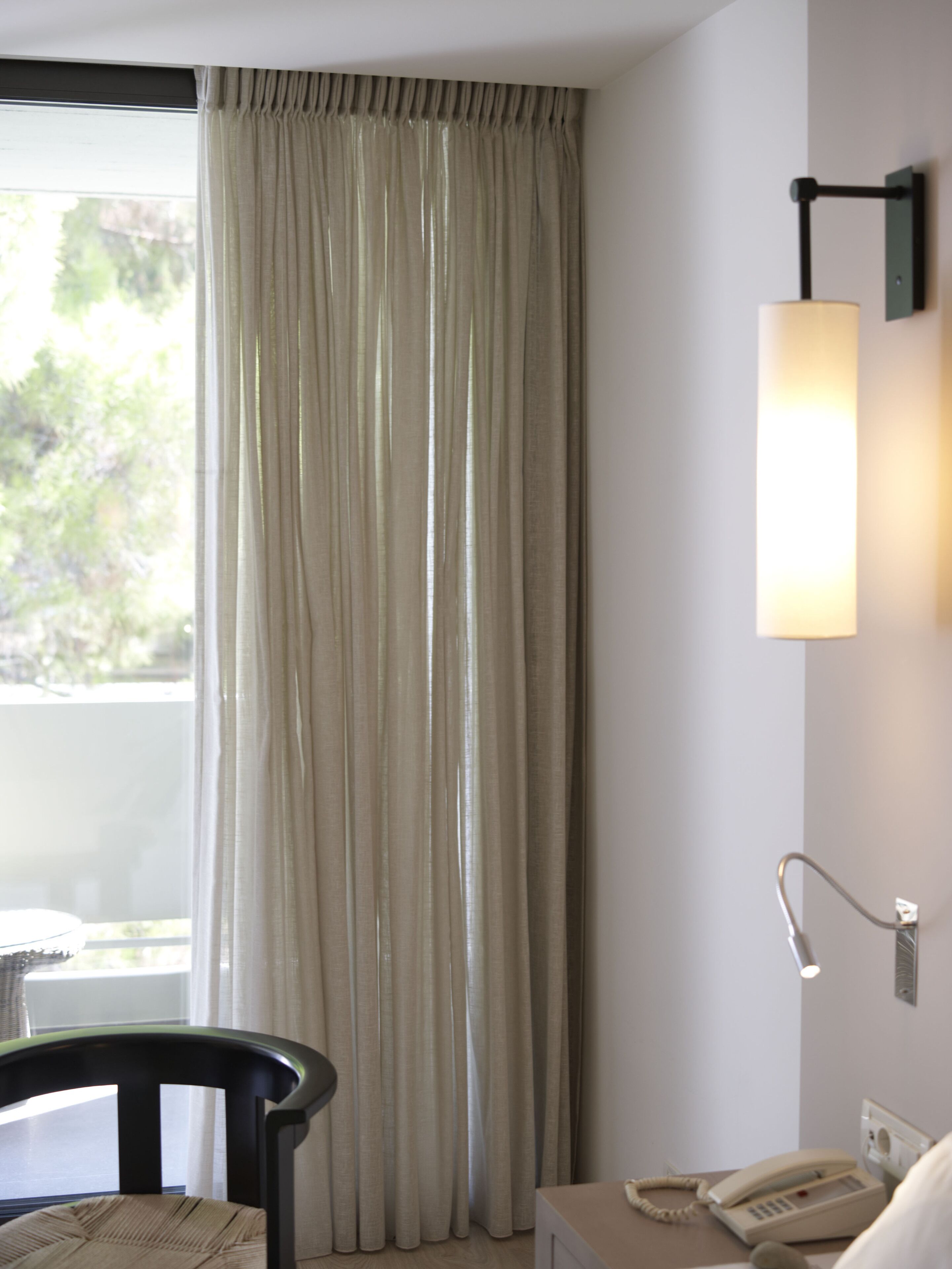 A hotel room with a large window, a chair, a bedside table with a lamp, and a telephone.