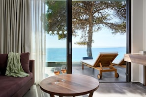 Un soggiorno moderno con un divano, un tavolo rotondo in legno con due bicchieri di vino, e vista sul mare tramite ampie vetrate.