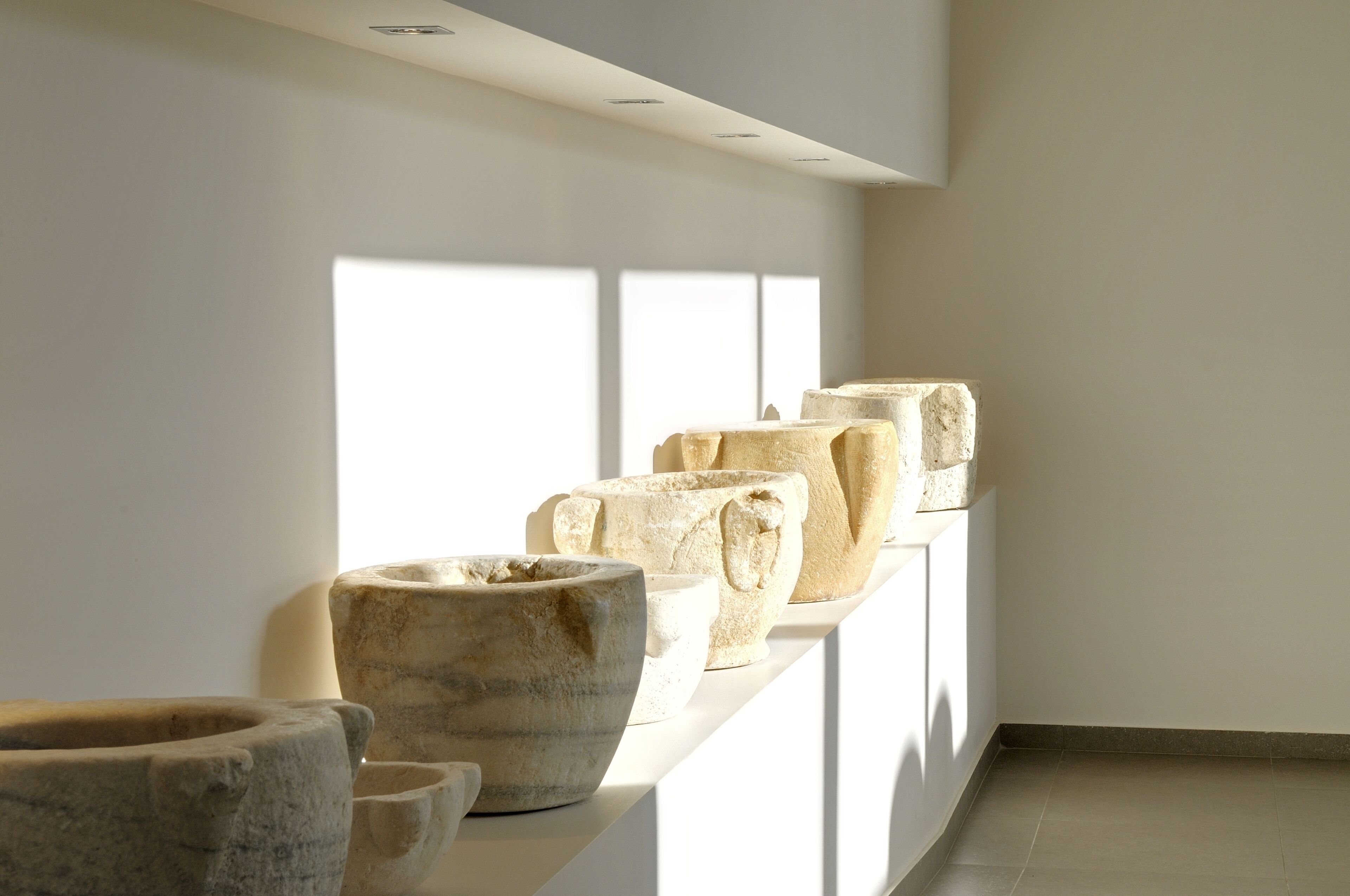 A series of stone vessels displayed on a white shelf in a well-lit room with large windows.