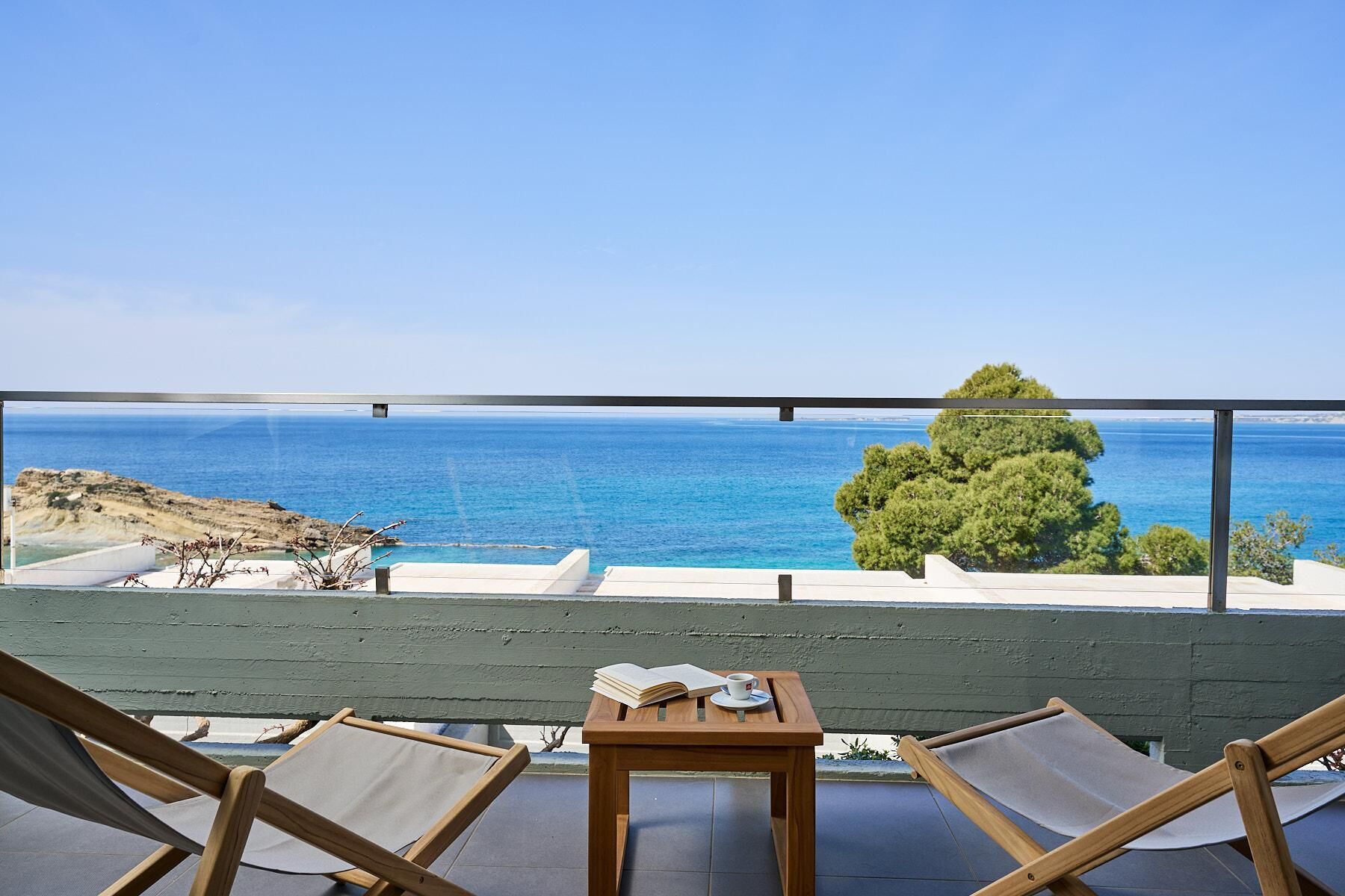 A balcony with two wooden chairs and a small table, overlooking a coastal view with clear blue sea and a tree.