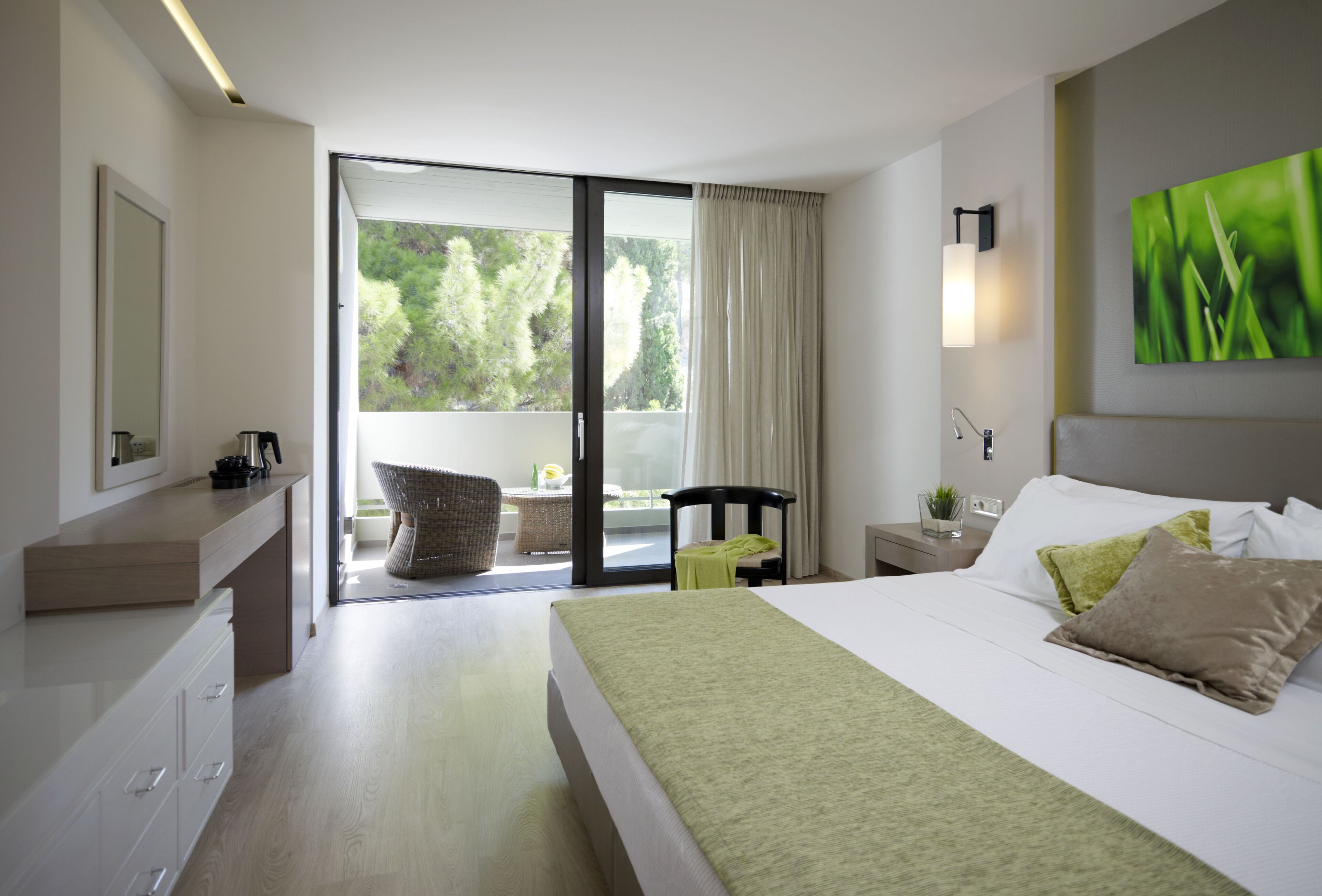 A modern hotel room with a large bed, a desk with a chair, a small table with a lamp, and a sliding glass door leading to a balcony with a view of trees.