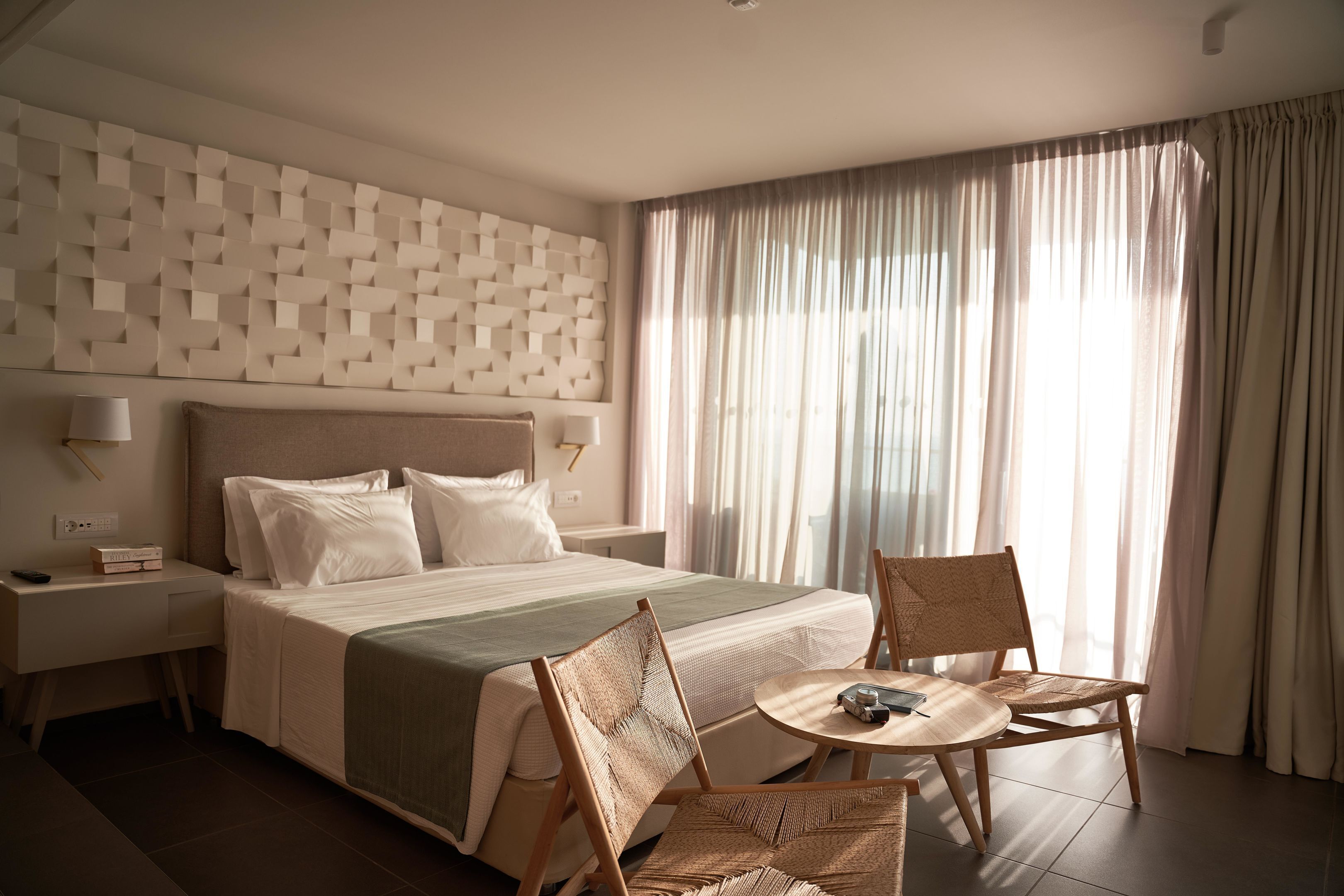 A hotel room with a large bed, two chairs, a small table, a nightstand, and a wall with a geometric pattern.