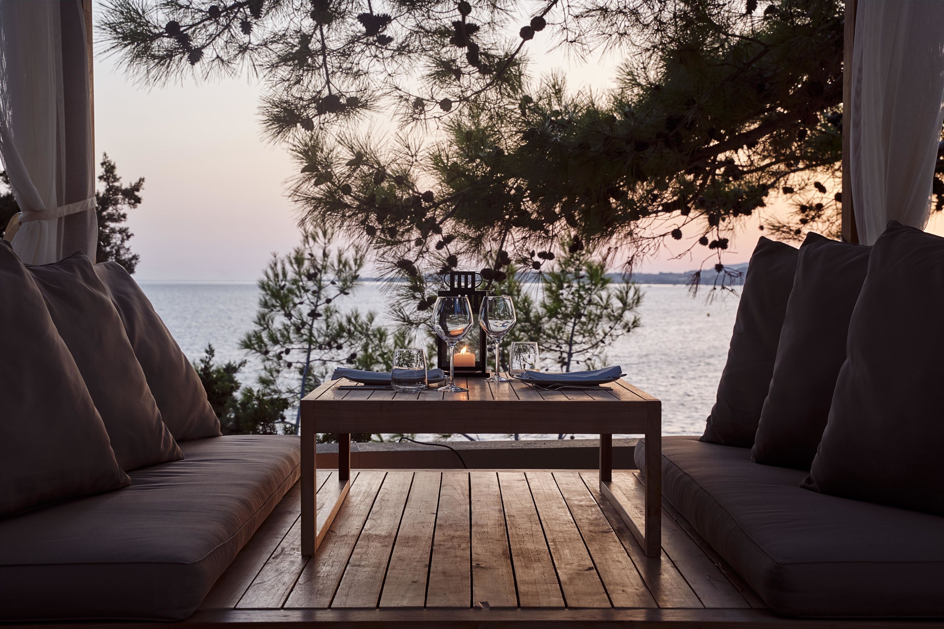 Una mesa de madera con vasos y una vela, frente a un atardecer sobre el agua, con un sofá y cortinas a cada lado.
