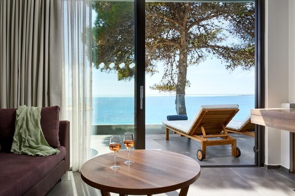 A modern living room with a sofa, a round wooden table with two glasses of wine, and a view of the sea through large glass doors. - White Rocks Hotel Kefalonia (Kefalonia)