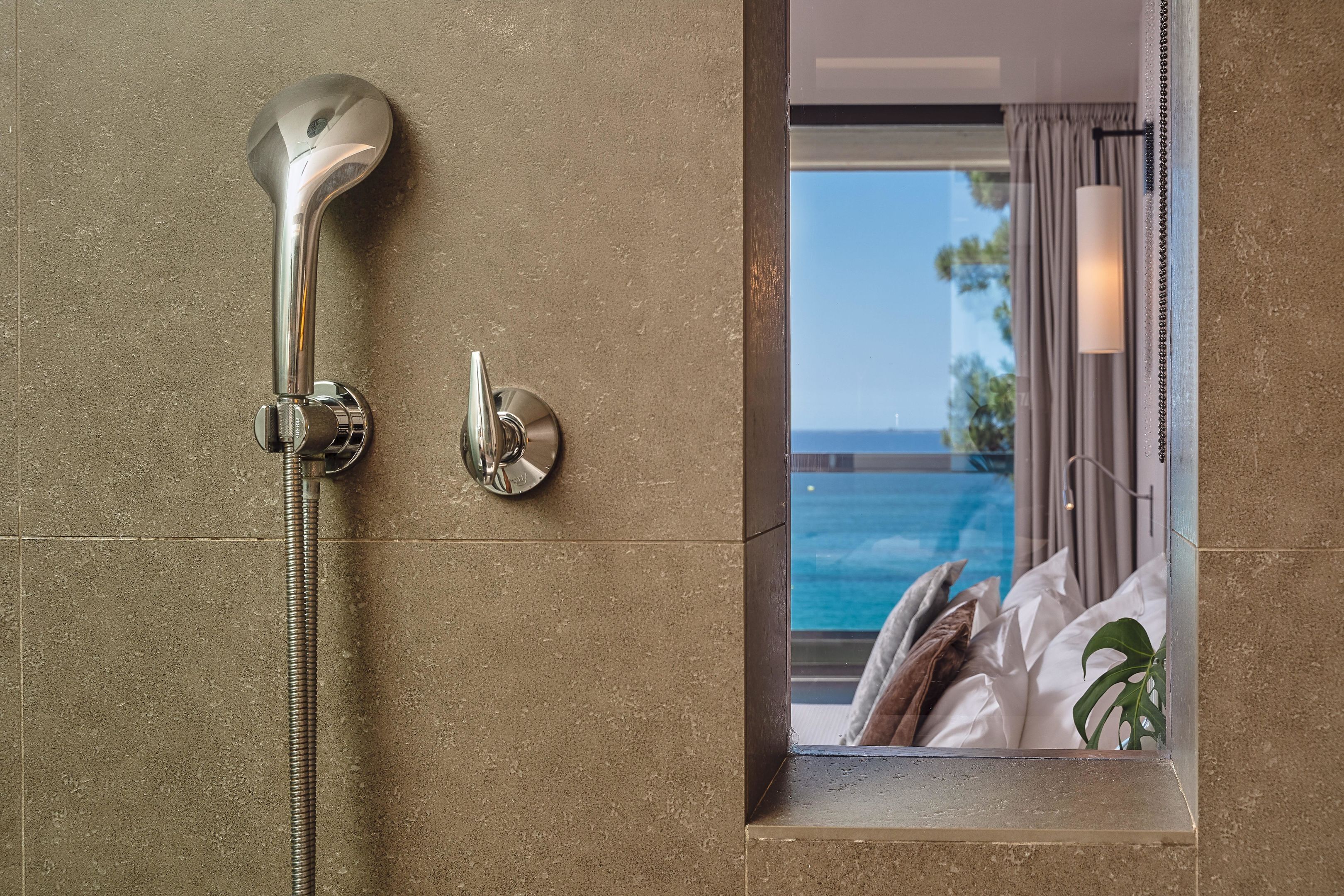 Une salle de bain avec douche, une vue sur une chambre avec un lit et une plante, et une piscine visible par la fenêtre.