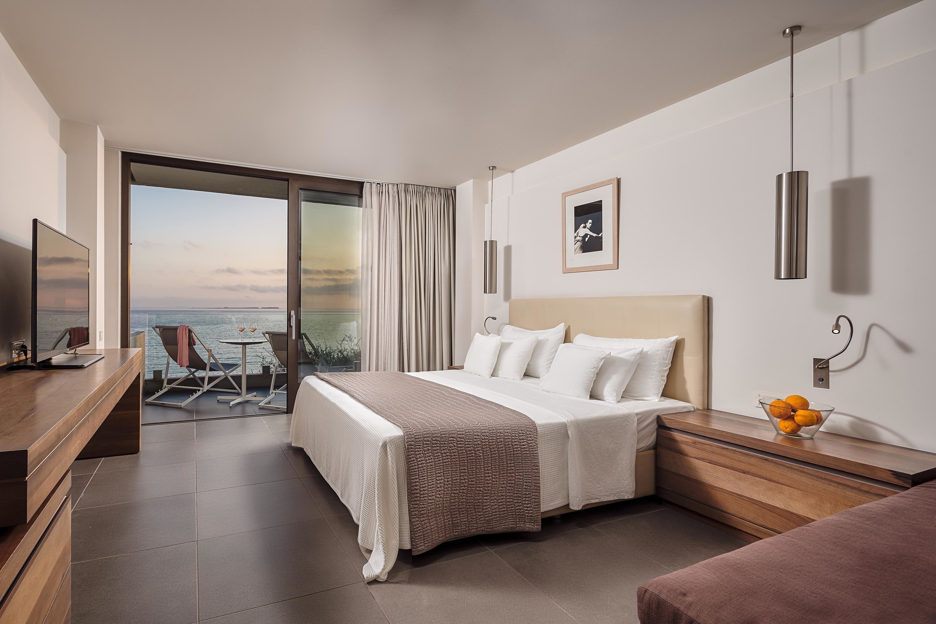 A modern hotel room with a bed, a desk, a chair, and a view of the sea.